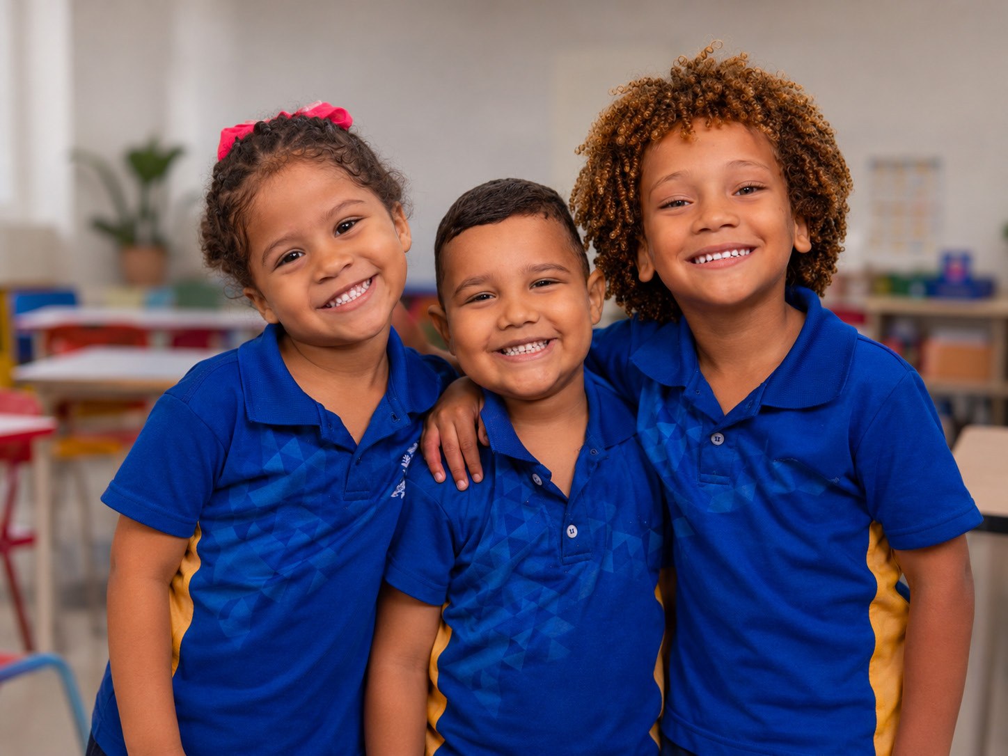 Três crianças da rede municipal sorrindo juntas, uniformizadas.
